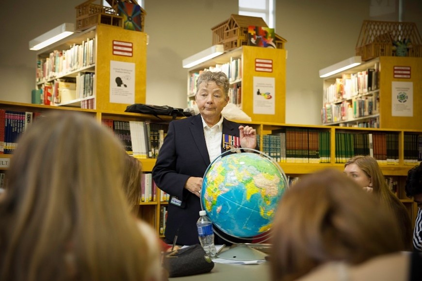 Une vétérane en uniforme s’adresse à des élèves dans une bibliothèque. Un globe terrestre coloré est posé sur la table entre les élèves..