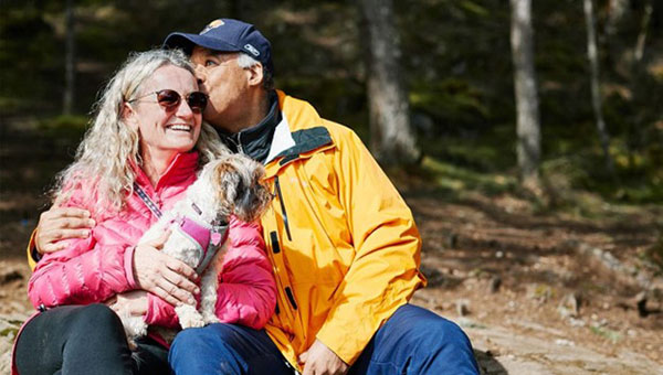 a smiling couple sitting in the woods with a small dog.