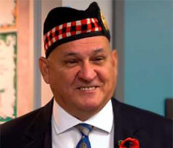 A smiling man wearing a black suit, blue patterned tie, red-and-black checkered hat, and ceremonial decorations. Medals and ribbons are on both sides of his jacket.