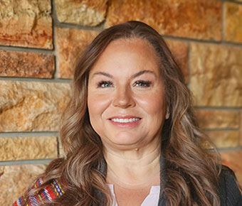 A smiling woman wearing a black pinstriped suit and colorful woven ceremonial sash. Multiple medals and pins decorate her sash and jacket.