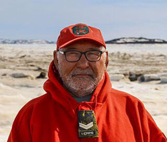 A bearded man wearing red cold-weather clothing, hood, cap, glasses, and an embroidered chest patch while standing on an icy shoreline.