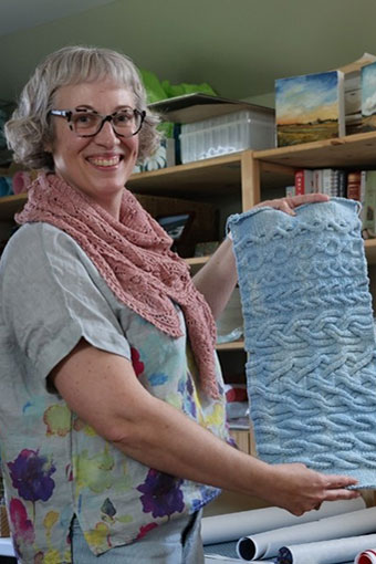 A smiling woman in craft-filled room holding large light-blue knitted piece.