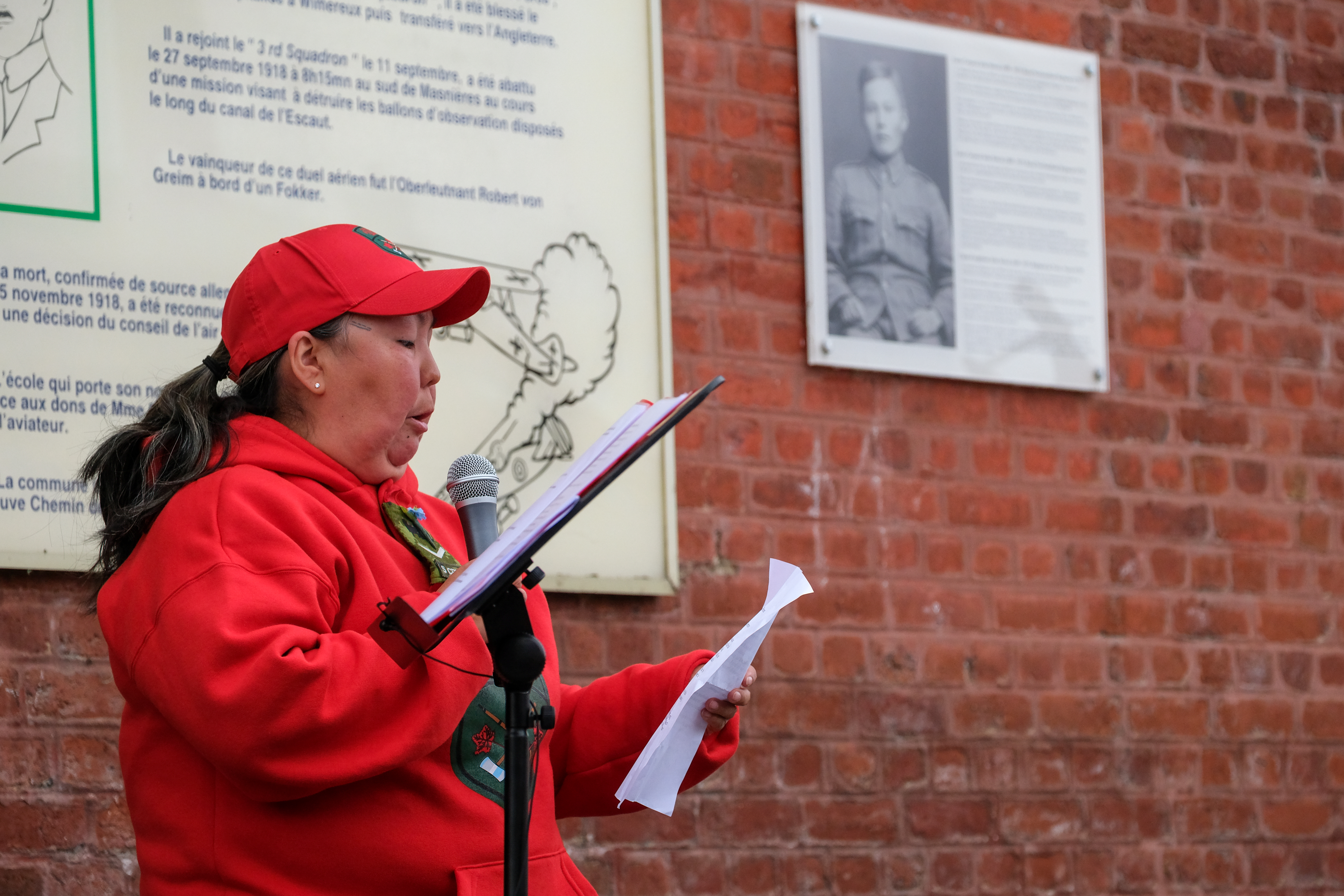 A person in a red jacket and cap reads from a sheet of paper into a microphone.
