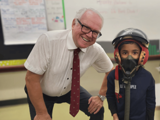 Photograph showing an older man in a white shirt and red tie smiling and crouching next to a young child wearing a pilot helmet and oxygen mask.