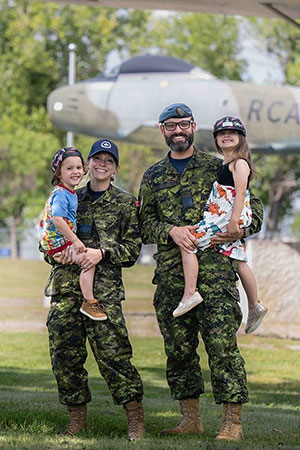 Deux personnes portant l’uniforme militaire camouflage sont debout, sur du gazon. Ils tiennent chacun un enfant dans leurs bras. Il y a un aéronef en arrière-plan.