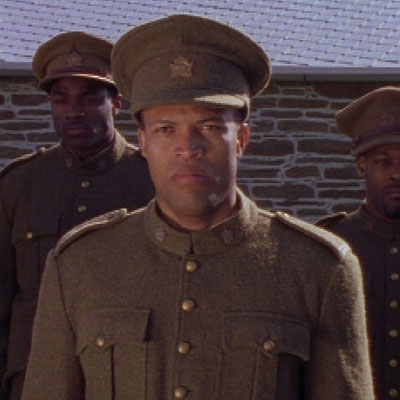 Une capture d’écran du film Honour Before Glory montre trois acteurs incarnant des membres du 2e Bataillon de construction, vêtus d’uniformes militaires de la Première Guerre mondiale, devant un bâtiment en pierre.