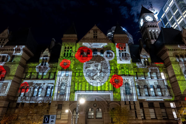 Une projection nocturne recouvre la façade d’un bâtiment historique de Toronto, en Ontario, affichant de grands coquelicots rouges et des images commémoratives illuminées sur toute sa façade.