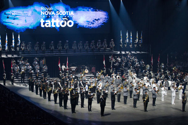 Un grand ensemble de musiciens et d’artistes se tient en formation sur le sol d’un aréna lors d’un événement de la Royal Nova Scotia International Tattoo Society, avec un fond de scène lumineux affichant le logo de l’événement et d’autres artistes positionnés sur une plateforme surélevée au-dessus.