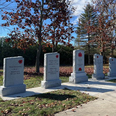 Dans la ville de Georgina, en Ontario, une rangée de cinq piliers commémoratifs en granit se dresse à l’extérieur, chacun gravé d’une feuille d’érable et d’inscriptions honorant différents groupes de militaires, avec des arbres et un ciel dégagé en arrière-plan.