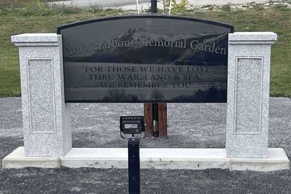 Le panneau du Jardin commémoratif de York Harbour, à Terre-Neuve-et-Labrador, est composé d’un panneau gravé sombre soutenu par deux piliers de pierre claire. On y lit l’inscription : « Pour ceux que nous avons perdus à la guerre, sur terre et en mer, nous nous souvenons de vous ».