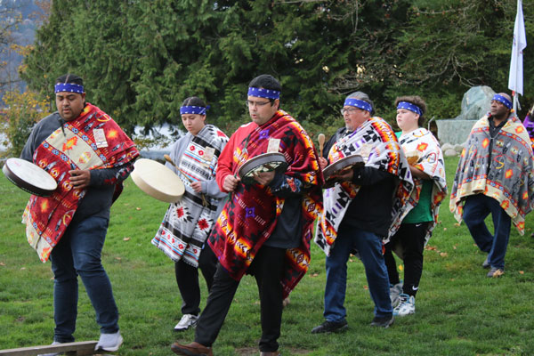 Lors d’un rassemblement de la nation Tsleil-Waututh, un groupe de participants vêtus de couvertures aux motifs colorés traversent une zone herbeuse en portant des tambours à main, avec des arbres et de l’eau visibles en arrière-plan.