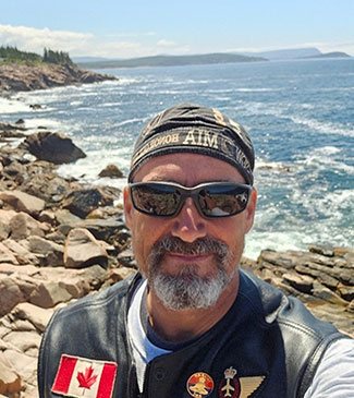Al McFarlane se tient sur le Cabot Trail, Cap-Breton, Nouvelle-Écosse, vêtu d’une veste de moto en cuir noir avec un drapeau canadien et des écussons militaires, de lunettes de soleil et d’un chapeau. Il sourit.