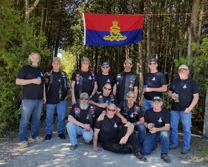 Un groupe de vétérans qui se nomment eux-mêmes le « Asshole Club » se tiennent debout à l’extérieur devant un groupe d’arbres; un drapeau militaire est hissé au-dessus d’eux.