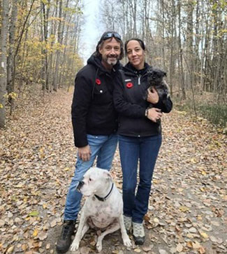 Al McFarlane se tient debout, le bras autour de sa compagne, sur un chemin de terre couvert de feuilles mortes. Elle tient un petit chien noir dans ses bras et le chien d’assistance de M. McFarlane, un boxer blanc, est assis à leurs pieds.