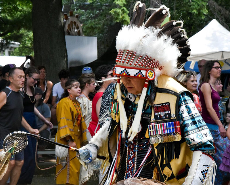 Major (Retd) Bob Crane wearing a headdress and full regalia, participates in a public gathering. He is holding a ceremonial staff and wearing multiple medals.