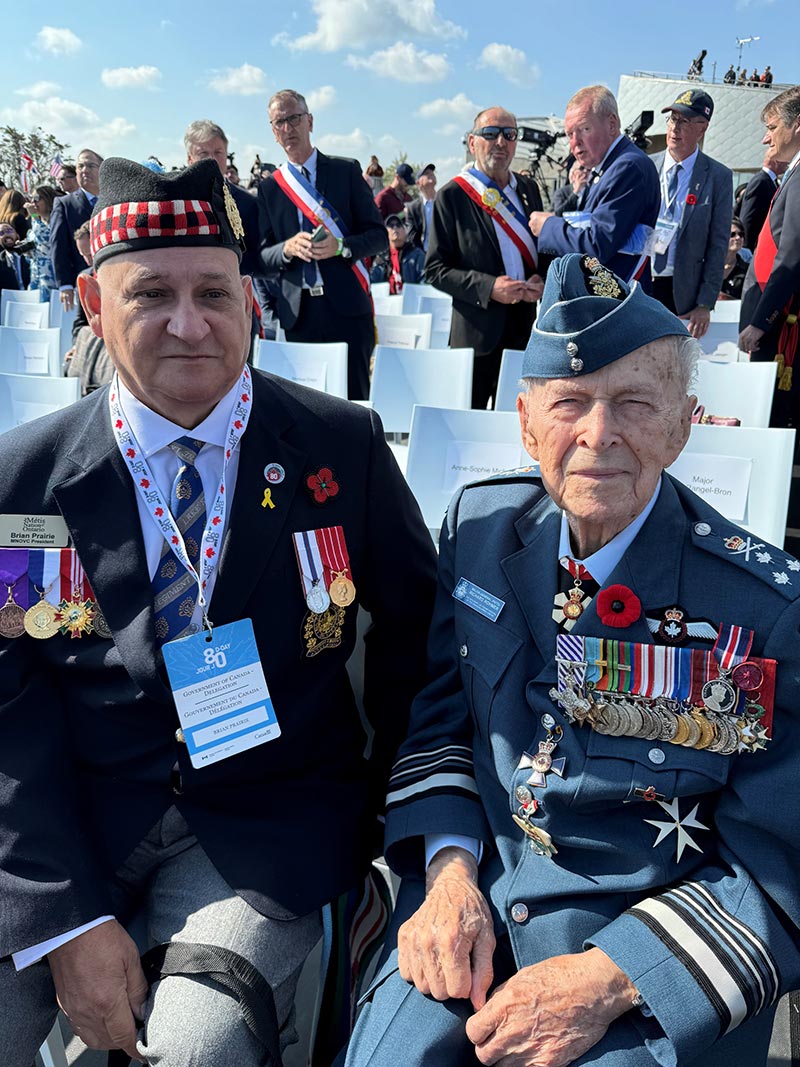 Brian Prairie sits in a white chair next to Honorary Lieutenant-General Richard Rohmer during a commemorative event at Juno Beach. There are people standing in the background.