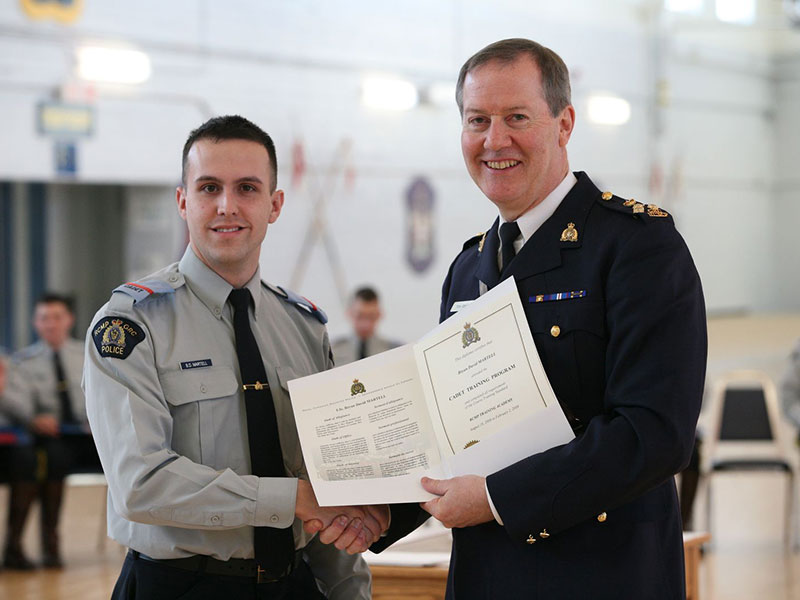 Bryan Martell is shaking hands with an officer. Both of them are wearing formal RCMP uniforms. The officer is holding Martell’s police cadet graduation certificate.