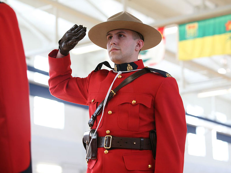 Bryan Martell is giving a salute in a red Mountie uniform and brown hat. He is standing in a white room.