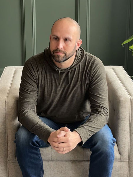Bryan Martell sitting on a chair with his hands clasped. He is wearing a sweater and jeans.
