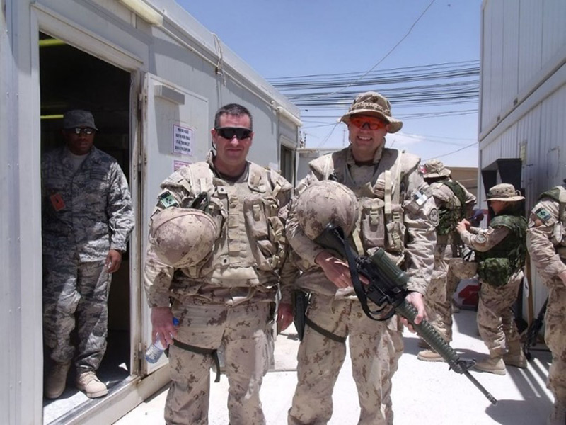 Carl Charbonneau, left, stands beside a fellow soldier on a military base in Afghanistan. The soldier beside him holds a rifle. They are both wearing grey-green camouflage gear and body armour and sunglasses and hold their helmets.