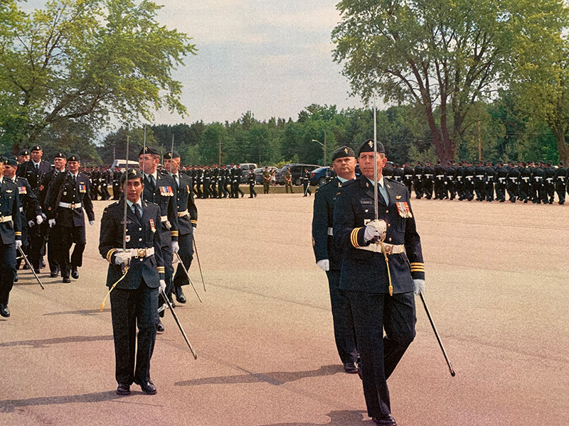 Paul Fuller, in military dress uniform, is leading a military parade. He is carrying a sword, in this right hand which is pointing to the sky. In his left hand is a baton. He is followed by a long line of Canadian Armed Forces members, all in dress uniform. They are parading on an asphalt area with large trees and blue sky behind them.