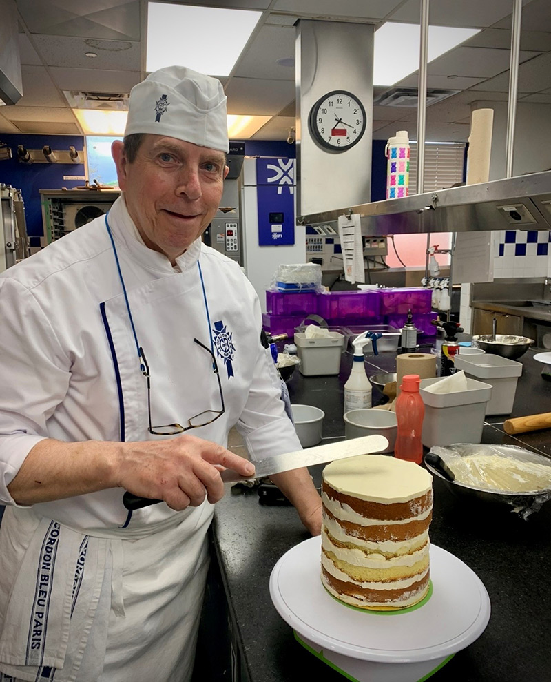 Paul Fuller, wearing white chef’s attire and hat, smiling at the camera. He is holding a flat spatula and is in the process of frosting a multi-layer cake. He is wearing a white chef’s coat and hat, a white apron and is standing in a commercial kitchen.