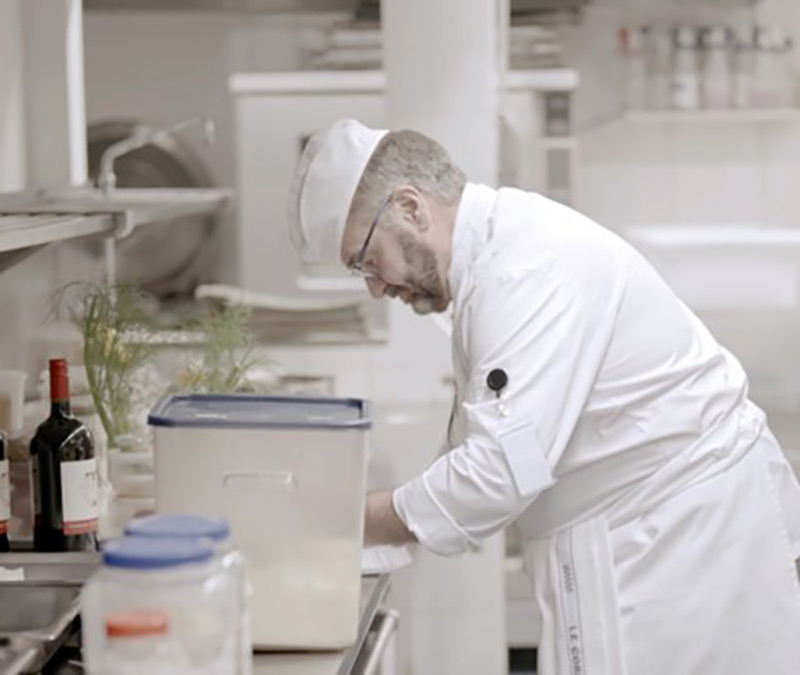 Carl Charbonneau, with graying hair and beard, leans over the counter in a commercial kitchen at Le Cordon Bleu Ottawa Culinary Arts Institute. In the foreground are a number of plastic containers with blue lids and a bottle of wine. He is wearing a white chef’s uniform and black rimmed glasses. 