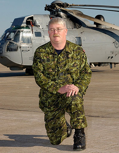 A middle aged Jack MacIsaac is on one knee, on concrete, in front of a military helicopter at 12 Wing Shearwater in Nova Scotia. He is wearing camouflage fatigues with black boots. His hair is short, he has a moustache and is wearing glasses.  