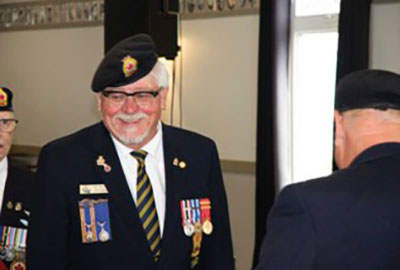 Older Veteran (Jack MacIsaac) in a formal portrait. He is smiling in to the camera and has a gray goatee and is wearing dark rimmed glasses. He is also wearing a blue suit jacket with medals on both sides of the jacket. He wearing a legion lapel pin as well as a beret with the legion emblem.