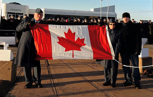 Three people holding a large Canadian flag in front of a large group of people in formal military uniforms. The man on the left (Jack MacIsaac) is holding the flag with black leather gloves, he is also wearing a long black trench coat, dark slacks, black dress boots and a dark coloured Airforce wedge cap. The woman on the right is wearing the same and the man beside her is wearing a dark coat, dark pants and a black toque.  