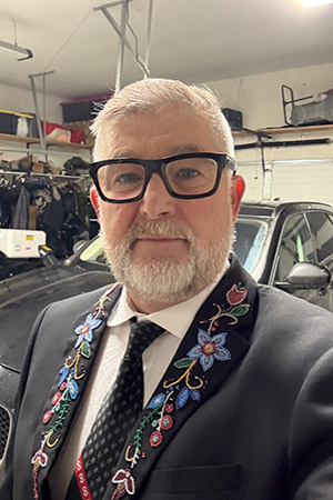 Chuck Isaacs stands in front of a black car in a garage. He is wearing large black glasses, white shirt and black tie and a black blazer the lapels of his jacket are beaded with traditional Métis blue and red flowers.