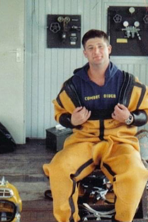 A young Chuck Isaacs sits in a yellow wetsuit, holding the front open to reveal the words “combat diver” on a blue hoodie underneath. A yellow diving helmet is on the ground beside him.