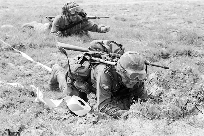 Chuck Isaacs and another soldier lie on the their stomachs in the grass. They are heavily armed and wearing fatigues and helmets and holding rifles.