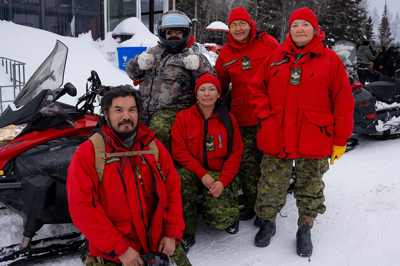 Cinq Rangers canadiens posent pour une photo devant une motoneige. Les quatre Rangers à l’avant portent les mêmes vestes et bonnets d’hiver rouges. L’homme assis sur la motoneige porte une veste de camouflage d’hiver et un casque, dont la visière est relevée. 