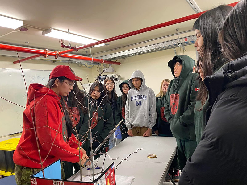 Une femme portant un chandail et une casquette rouges (Chastity Koosees) montre les compétences des Rangers à une classe de Rangers juniors. Un groupe d’environ 10 adolescents se tient autour d’une table et observe ce que fait Koosees. 