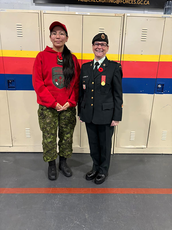 Deux femmes posent devant une rangée de casiers. Il y a des lignes horizontales jaunes, rouges et bleues qui traversent le centre des casiers. La femme à gauche (Koosees) porte un chandail rouge, une casquette rouge et un pantalon de camouflage, les mains jointes devant elle. La femme à droite (Butt) porte un uniforme militaire des FAC. Les deux regardent directement la caméra en souriant. 