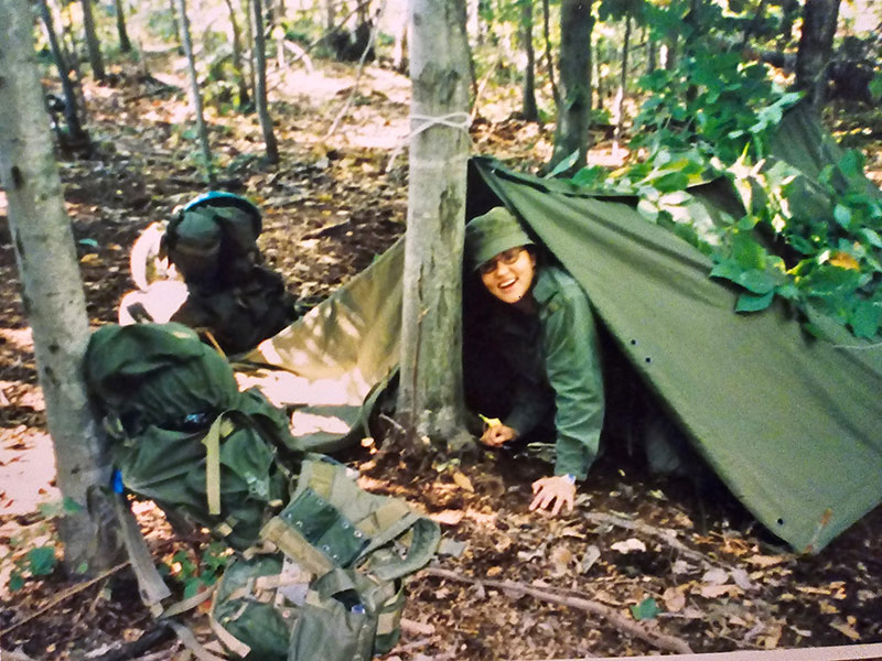 Plan large d’une zone boisée. Cheryl sort la tête d’une tente verte. Elle porte une tenue militaire verte. Des sacs à dos sont posés contre les arbres autour de la tente.