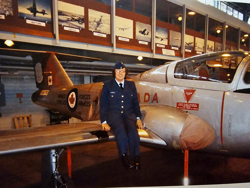 Plan large de Cheryl assise sur l’aile d’un avion des Forces canadiennes. Elle porte un uniforme militaire bleu et un calot. Elle sourit à la caméra.