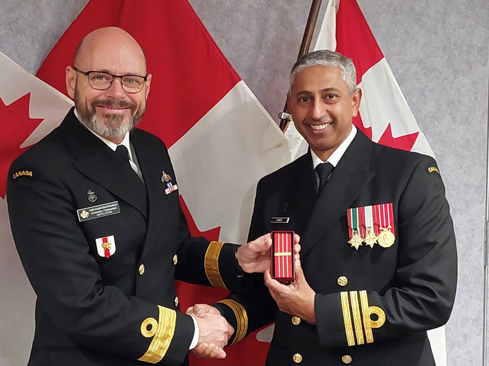 Two men in military dress uniforms pose for a photo in front of three Canadian flags. The man on the left is presenting the one on the right with a medal.
