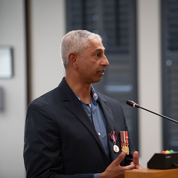Desmond James, wearing a blue dress shirt and navy blazer with military medals speaks at a microphone.