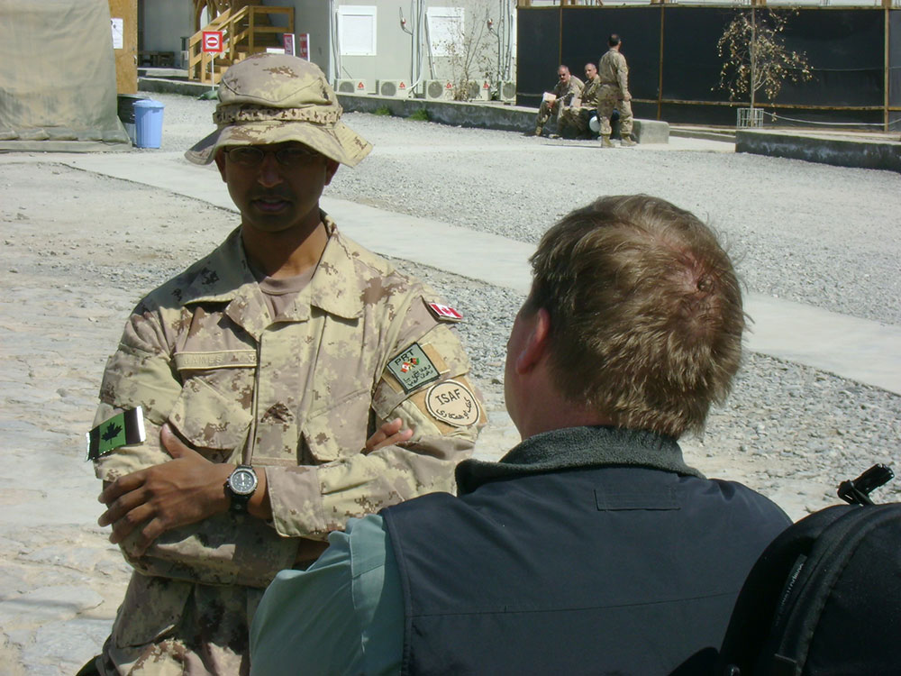 A man (Desmond James) in military fatigues stands with his arms crossed speaking to another man whose back is to the camera.