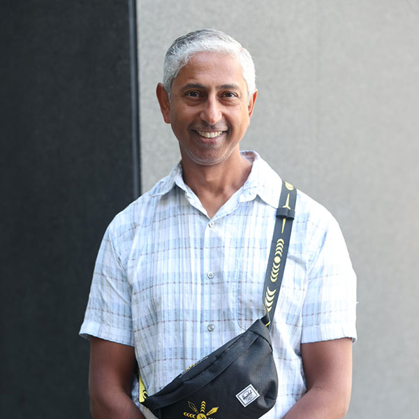 Desmond James stands smiling at the camera. He is wearing blue jeans and a short-sleeved white shirt with a black cross-body bag across his chest. His hands are clasped in front.