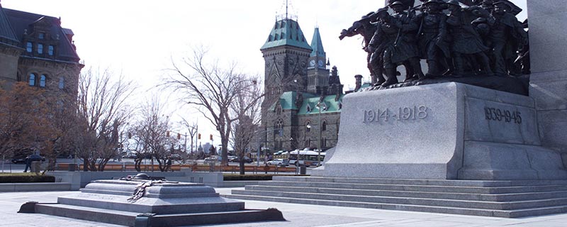 A sarcophagus made of stone sits at the foot a statue with metal soldiers standing atop a base that has the dates of the World Wars on it. Behind this National War Memorial are the green-roofed Canadian Parliament buildings.