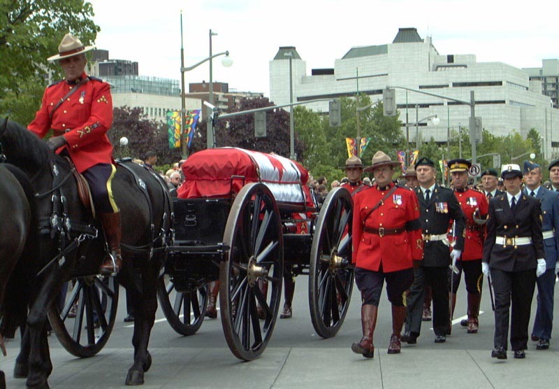 A casket draped with a Canadian flag sits on a wagon pulled by a black horse with a Royal Canadian Mounted Police officer atop through a city street. Another RCMP officer walks beside the casket, as do members of the Armed Forces in ceremonial dress uniforms.