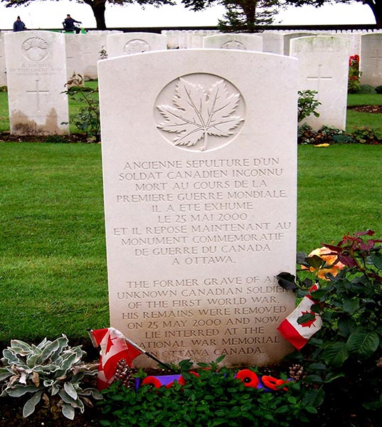 A white gravestone with a maple leaf and text sits behind plants and small Canadian flags. The text is in French and then English, explaining why the grave is empty, as the body now sits in the Tomb of the Unknown Soldier in Ottawa.