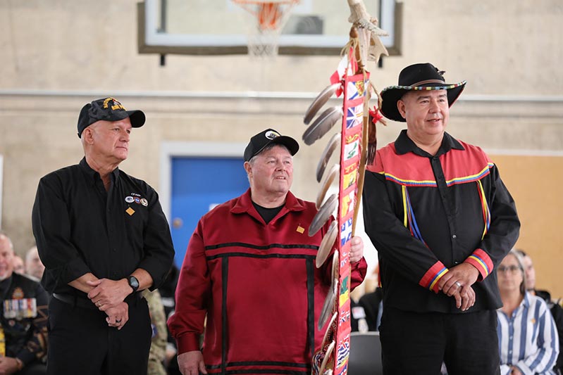 Trois hommes se tiennent debout derrière un bâton à exploits cérémoniel orné de plumes d’aigle et comportant un drapeau représentant chaque province et territoire du Canada. Tous les trois portent un chapeau, et l’homme à droite arbore des rubans autochtones sur sa chemise noire.