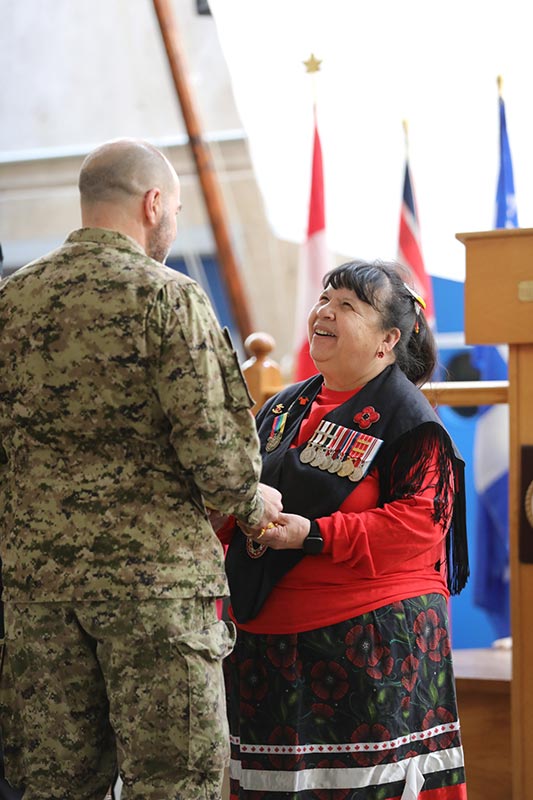 Une femme des Premières Nations, vêtue d’une veste noire ornée d’un coquelicot en perles et de médailles militaires, ainsi que d’une jupe à rubans traditionnelle, est debout, souriante, et tient la main d’un homme en tenue militaire. Ce dernier tourne le dos à la caméra.