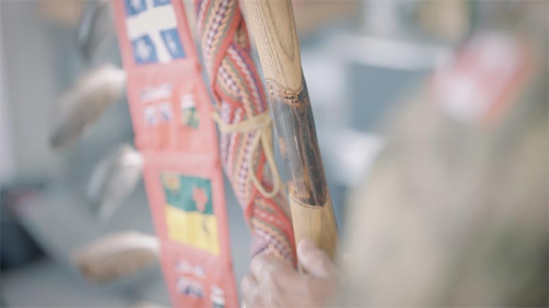 Gros plan sur le bâton à exploits. Une main tient le bois, au-dessus de la main se trouve une tache sombre dans le bois en forme de cœur, entourée par deux autres cœurs. Une ceinture métisse multicolore enveloppe l’autre partie du bâton. Les drapeaux représentant chaque province canadienne sont placés dans un ruban à l’extérieur du bâton. Les drapeaux sont ornés de plumes d’aigle.