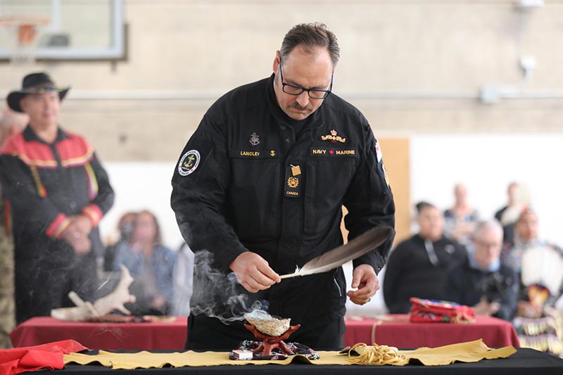 Un homme en uniforme de la Marine tient une grande plume d’aigle tandis que de la fumée s’élève d’un coquillage.