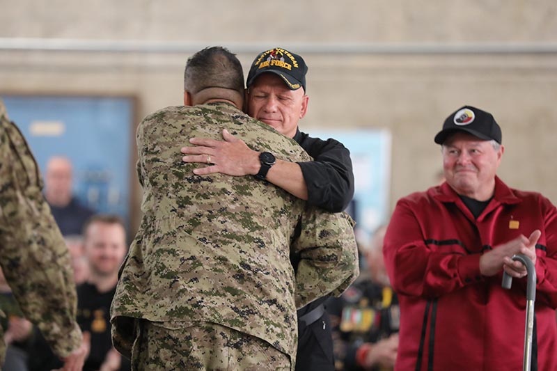 Un homme en tenue militaire serre dans ses bras un homme plus âgé vêtu d’une chemise noire, d’une casquette noire de la Force aérienne et d’une montre noire. Les yeux de l’homme plus âgé sont fermés tandis qu’un autre homme, une canne à la main, les observe.
